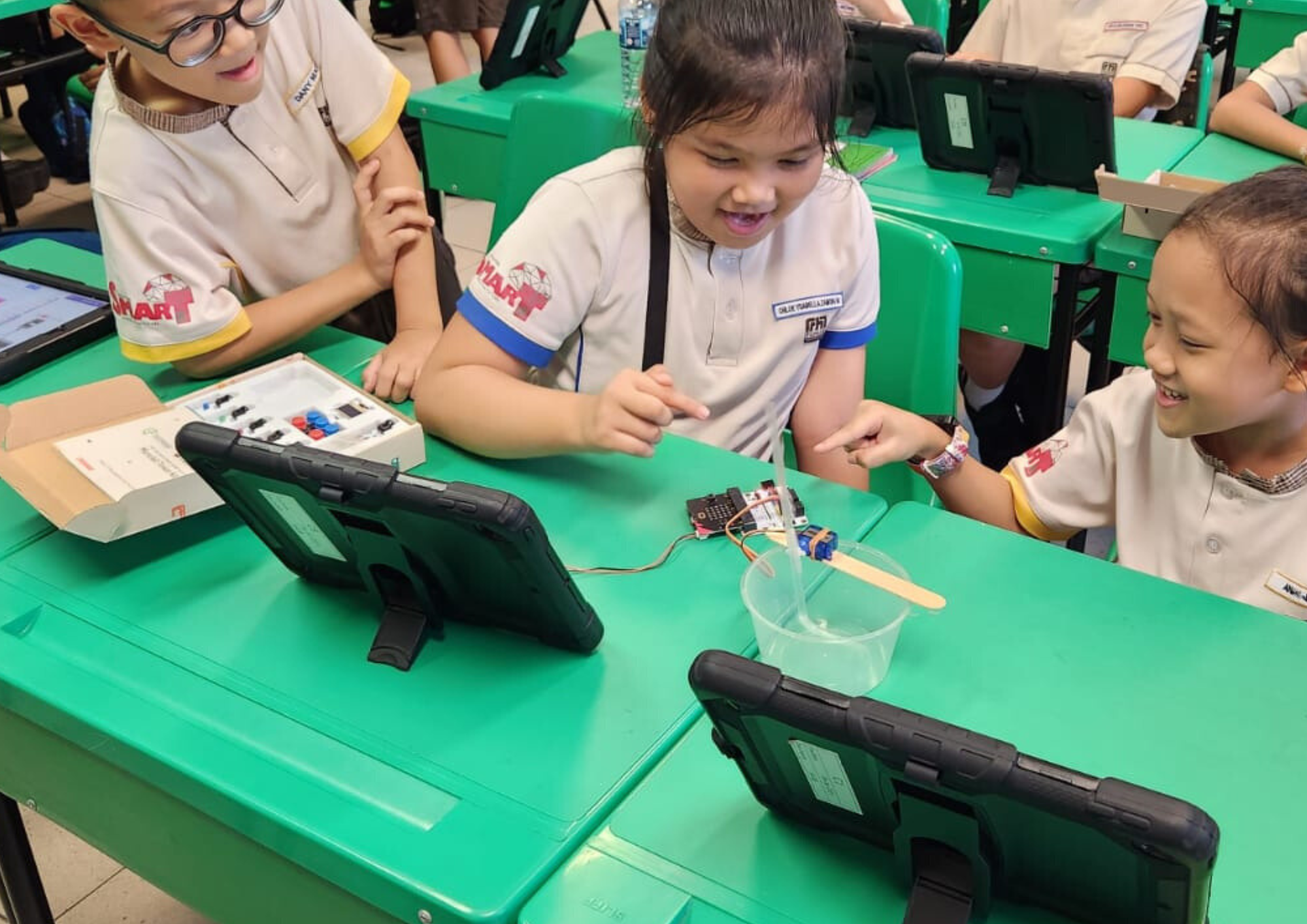 Three children in school uniforms smile while working on a small electronics project at green desks with tablets.